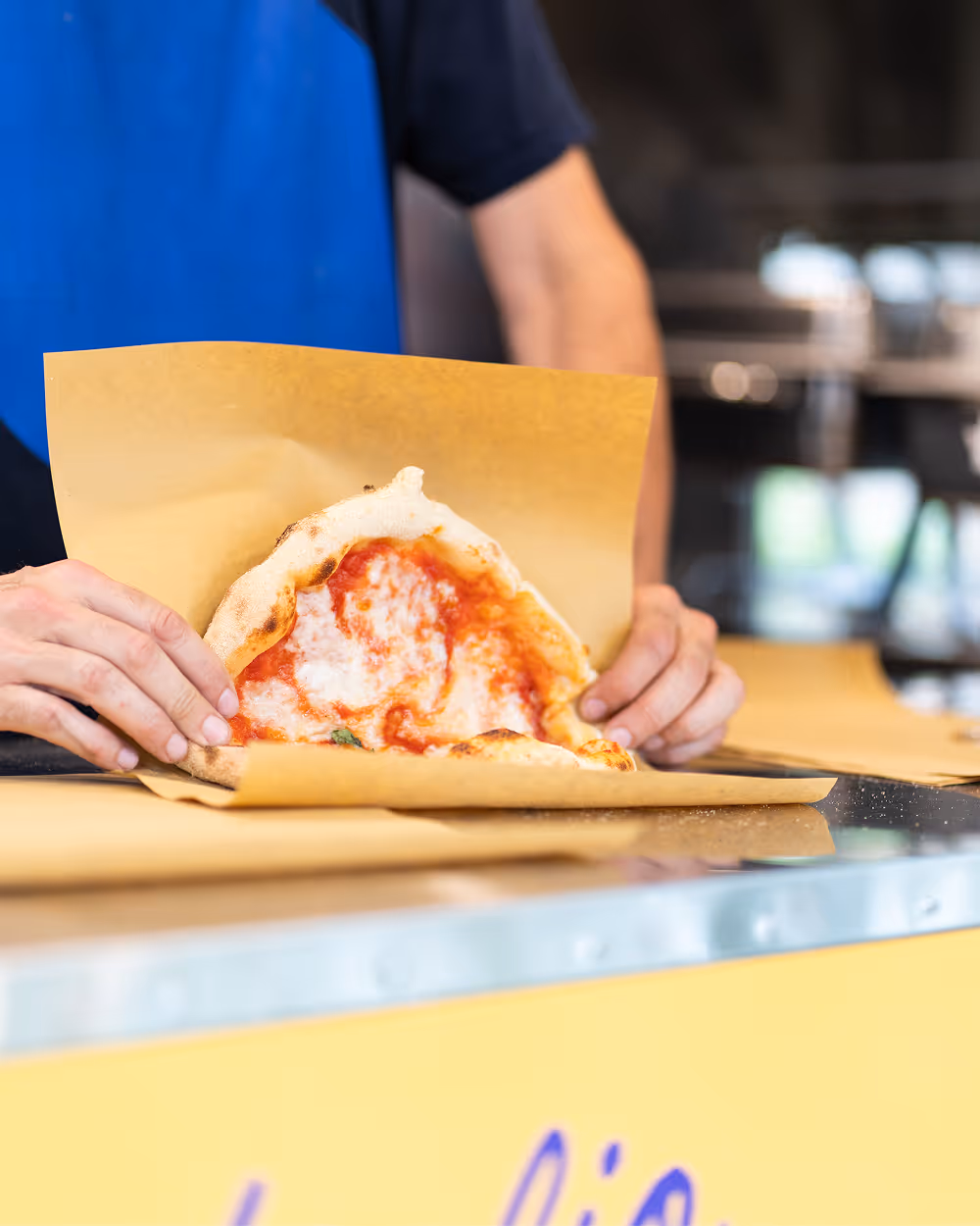 Person holding a folded pizza slice with tomato sauce and melted cheese on brown paper.