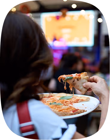 Person holding a pizza slice with melted cheese on a white plate in a blurred indoor setting.