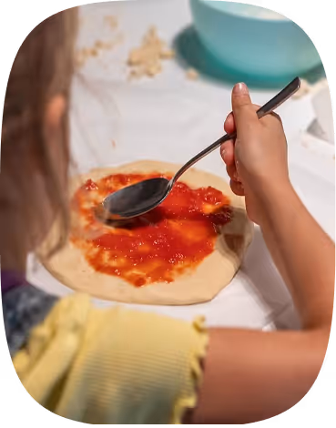 Child spreading tomato sauce on pizza dough with a spoon on a white surface.