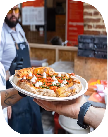 Hand holding a plate with a fresh pizza topped with dollops of creamy cheese and herbs in a restaurant setting.