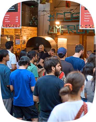 Crowd of people waiting indoors near a wood-fired pizza oven with menu boards above.