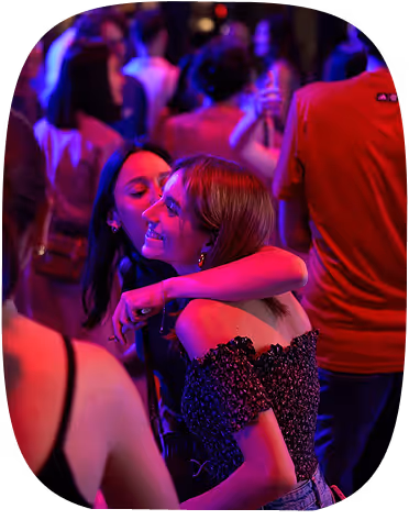 Two women smiling and hugging in a dimly lit party crowd with colorful lights.