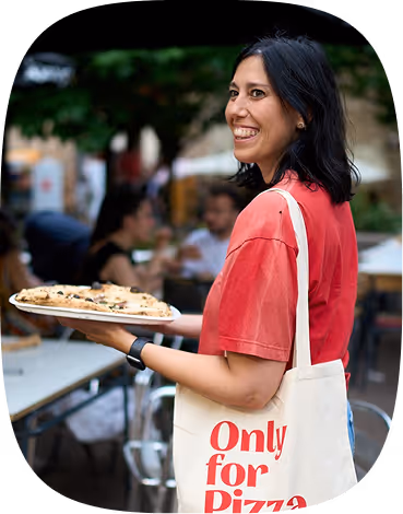 Smiling woman in red shirt holding pizza with tote bag that says 'Only for Pizza' outdoors.