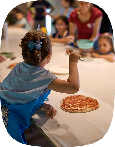 Child with blue hair bow and apron holds spoon over a pizza on a long table with people watching.